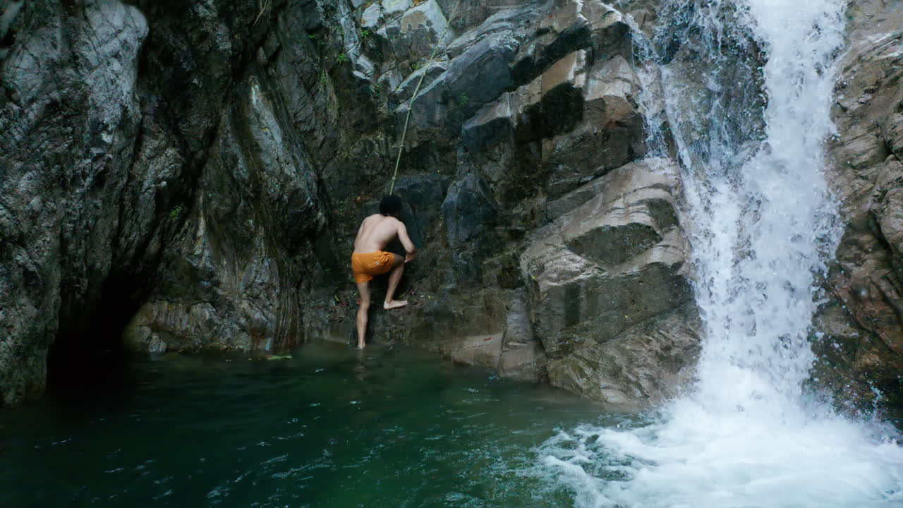 Uomo che esce dall'acqua per arrampicarsi con la corda sulla scogliera rocciosa della cascata, dall'alto