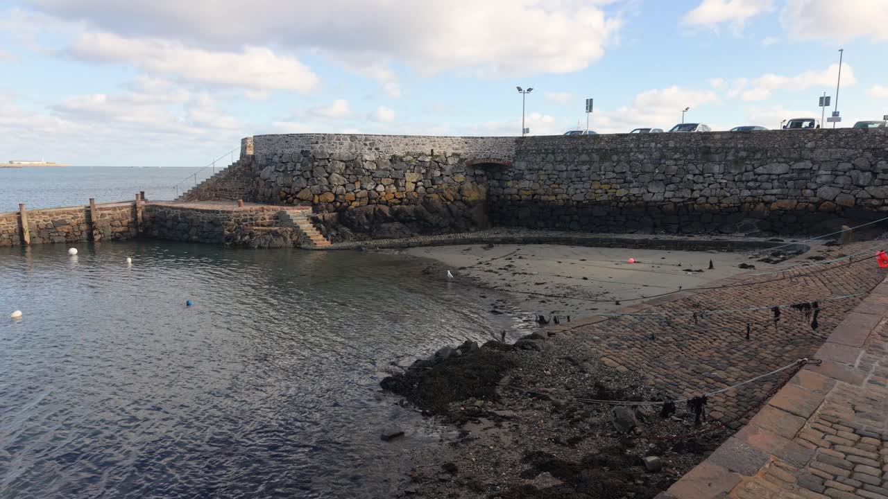 Riprese con drone di Salerie Corner a St Peter Port, Guernsey, con l'acqua che entra dolcemente nell'antico porto a metà marea, una piccola barca all'ancora sotto il sole dorato, acqua limpida e calma e vedute verso St Sampson