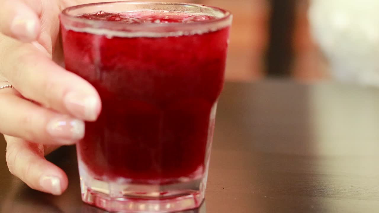 A hand twirls and stirs a red beverage in a glass, set on a table in a well-lit environment