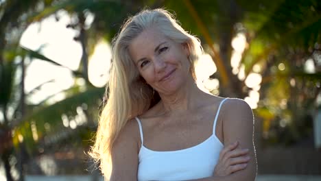 Closeup-intimate-portrait-of-backlit,-pretty-mature-woman-as-she-smiles-at-camera-with-a-palm-tree-behind-her