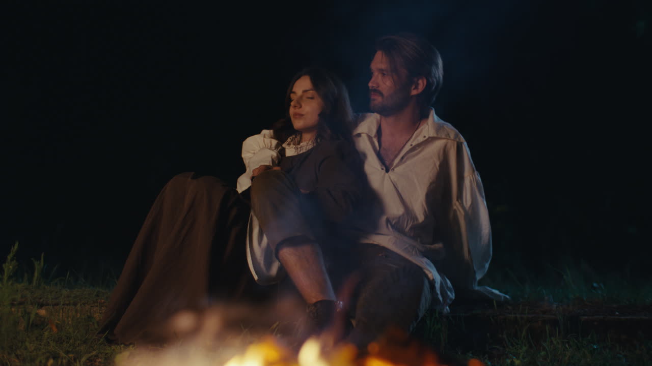 Couple in Period Clothing Sitting by a Campfire at Night