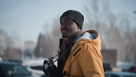 side view freelance photographer strolling snowy city street scanning surroundings through camera held at waist level, wearing yellow parka and black beanie amid parked cars, trees and sky