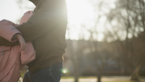 a little girl in a pink cap and jacket being turned around playfully by a man wearing a brown jacket and blue jeans. the scene captures a joyful moment with warm sunlight filtering through