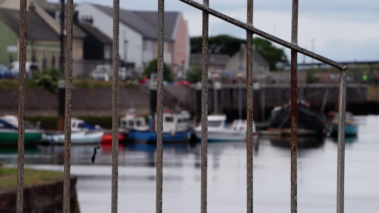 Rusty metal railings frame a soft-focus view of small fishing boats docked in a peaceful harbor, with calm water and houses in the background
