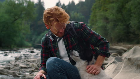 traveler petting dog at river shore. redhead man spending time with pet friend