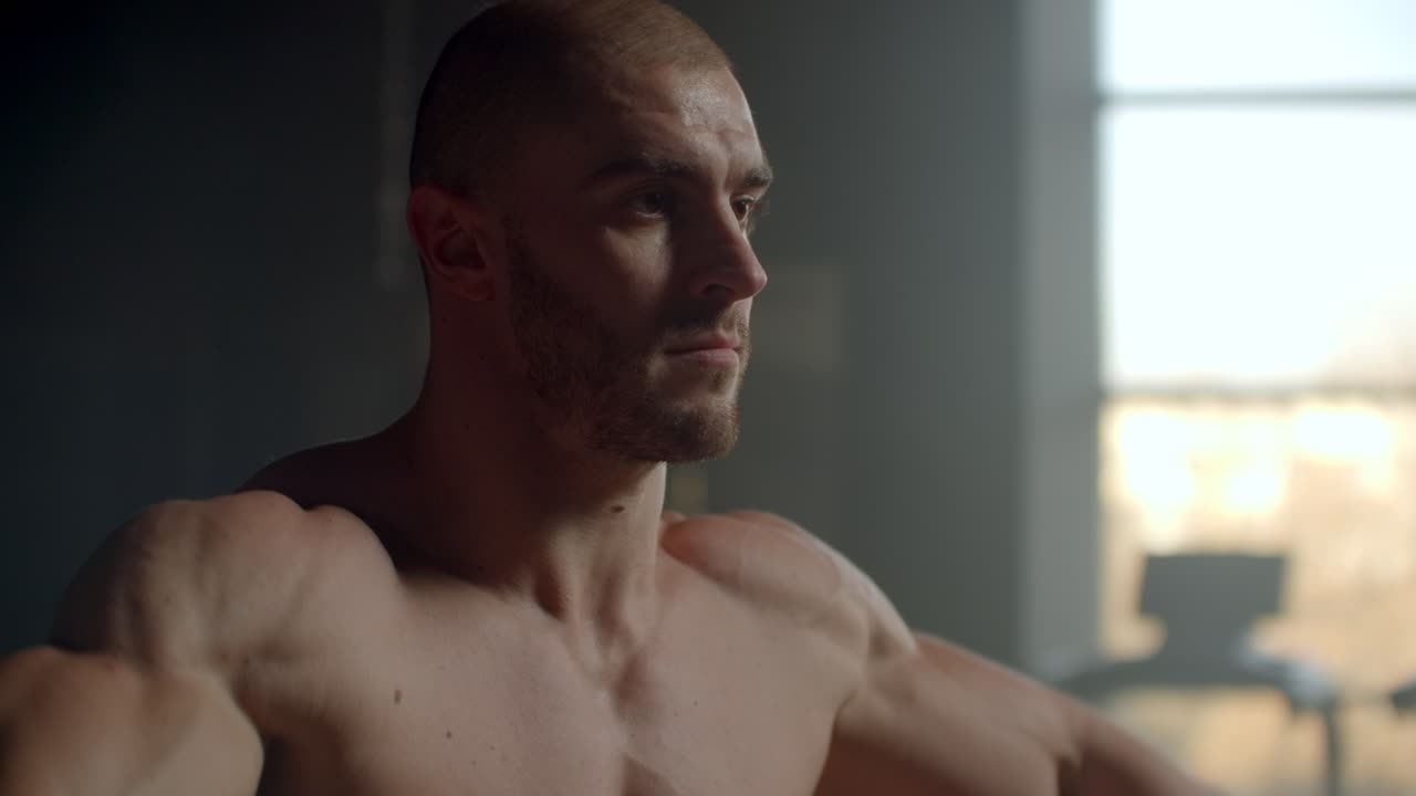 티셔츠를 입지 않은 남자가 창문 앞에서 덤벨을 들어올려 어깨 근육을 위한 운동을 하고 있습니다.건강한 생활 방식과 근육 질량을 얻기 위한 운동.강한 의지를 가진 운동선수의 근력 훈련.