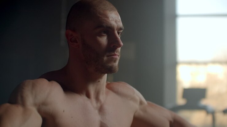 A man without a t-shirt lifts dumbbells in front of the window doing exercises for the shoulder muscles bodybuilder training. Healthy lifestyle and exercises to gain muscle mass. Strength training of a strong willed athlete.