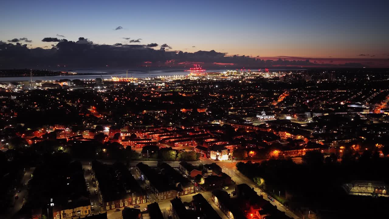 Drone descending over Liverpool at dusk, revealing warm-lit homes, red-lit cranes, and wind turbines by the water—showcasing the city’s mix of community, history, and modern energy