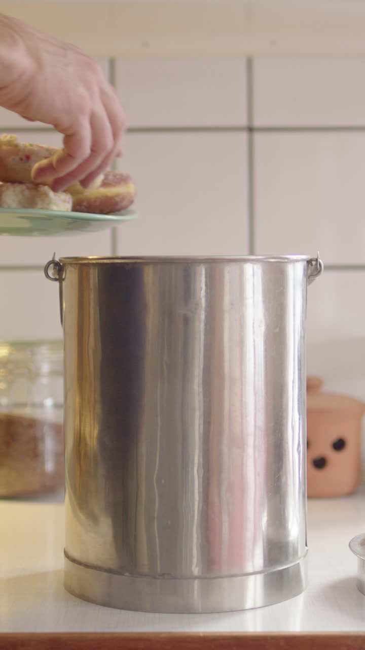 Man dumps stale buns and donuts into metal food waste bin in kitchen, vertical
