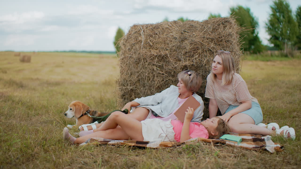 강아지와 함께 가족 휴식, 작은 소녀가 책을 읽고, 근처에 초록색 책, 어머니와 할머니가 광활한 시골에서 풀에 대항하여 휴식을 취하고 있습니다.