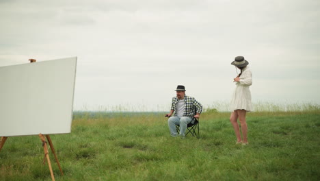 woman in a white dress stands with her hands on her hips, observing as an artist in a checked shirt demonstrates a sitting pose on a chair in a grassy field. an easel with a board is positioned nearby