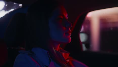 close up portrait of a female commuting home in a backseat of a taxi at night. beautiful woman passenger looking out of window while in a car in city street with working neon signs. cinematic footage.