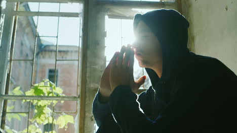 Teenager-In-The-Hood-Sits-On-A-Window-Sill-Holding-His-Hands-In-His-Head-To-Pray-In-The-Rays-Of-The-