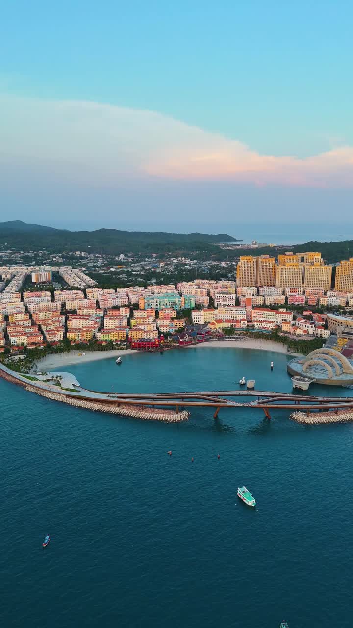 Vertical aerial view of the sunset over Phu Quoc Island, Vietnam, highlighting a replica Italian-style town. The colorful buildings along the coastline contrast with the serene waters and hills.