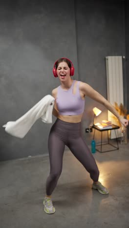 vertical video a happy and joyful brunette girl in a purple sports uniform with red wireless headphones dances and has fun holding a white towel in her hands after her exercise in a modern apartment at home