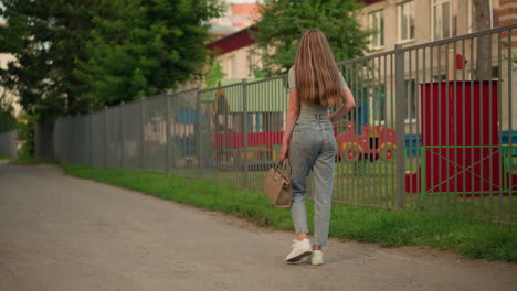 vista trasera de una mujer caminando por un camino al lado de una valla de hierro, sosteniendo un bolso de mano beige, su largo cabello rubio fluye por la espalda mientras camina con estructuras de patio de recreo visibles detrás de la valla