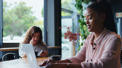 young businesswoman with coffee working on laptop sitting in coffee shop with colleague in background