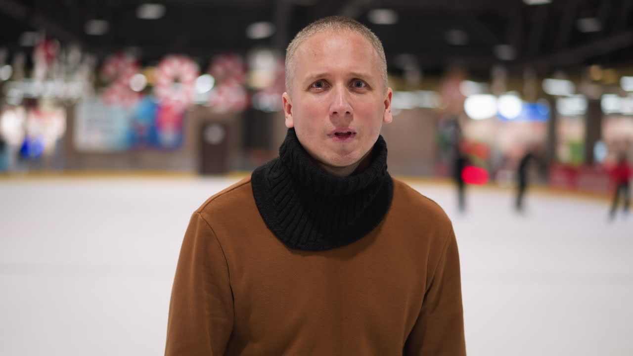 갈색 스웨터와 검은 스카프를 입은 중년 발머리 남자가 실내 링크의 배경에서 얼음 스케이터의 흐릿한 시각으로 카메라에 미소 짓고 있습니다.