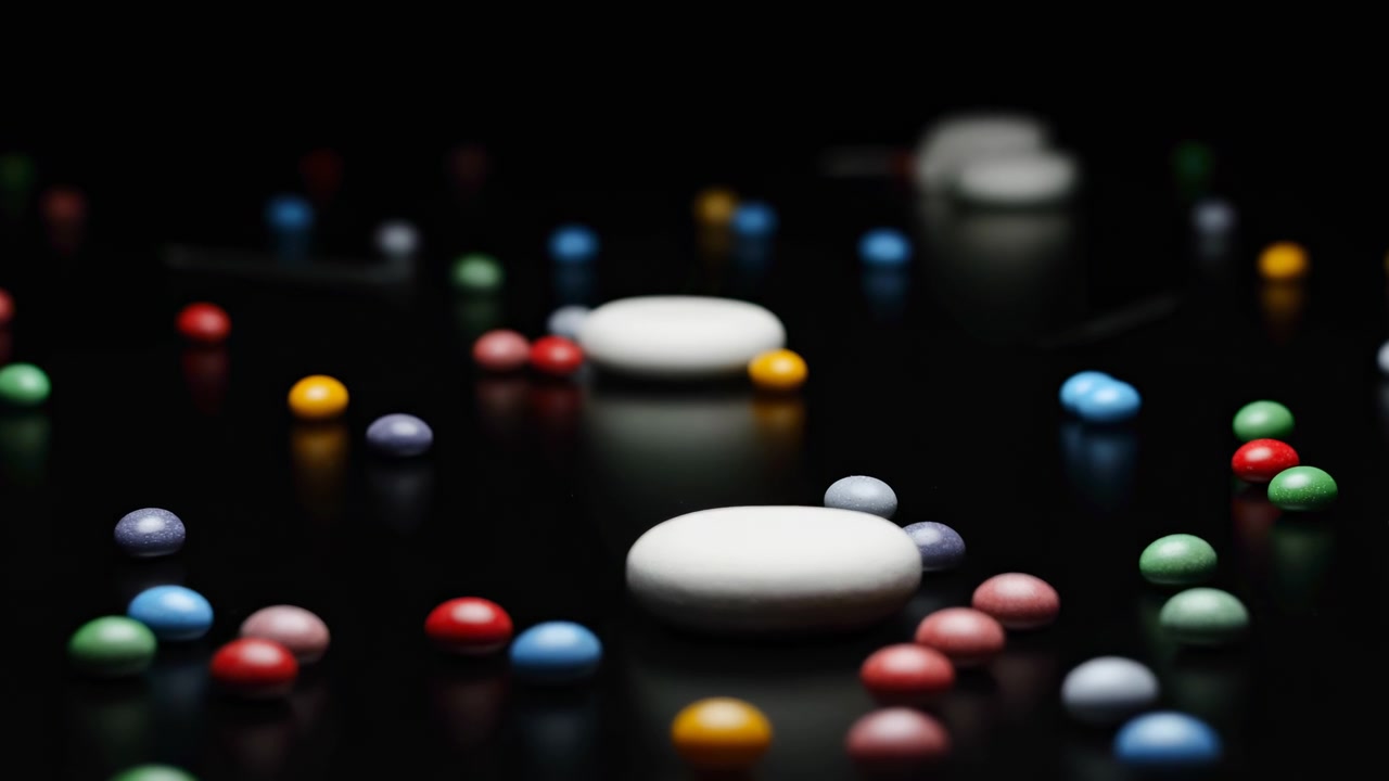Pilules colorées et deux gélules blanches éparpillées sur une surface noire brillante avec de fortes réflexions, éclairage de studio en basse lumière, faible profondeur de champ et profondeur visuelle subtile pour une image pharmaceutique abstraite.