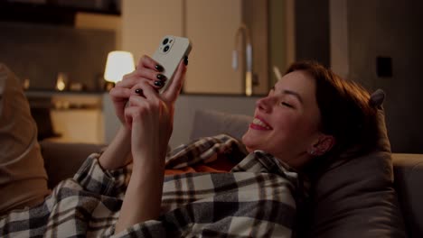 A-happy-and-confident-brunette-girl-in-a-plaid-shirt-lies-on-a-gray-sofa-and-types-on-social-networks-using-her-white-smartphone-in-the-evening-in-a-modern-apartment