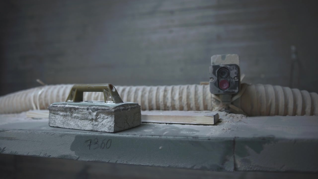 Close-up view shows plaster float resting on dusty workbench beside plaster block, with corrugated extraction hose, switch box, painted wood slats, and worn metal details in muted industrial light