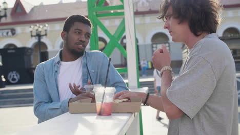 male friends chatting while eating pizza and drinking, standing at an outdoor table in the street