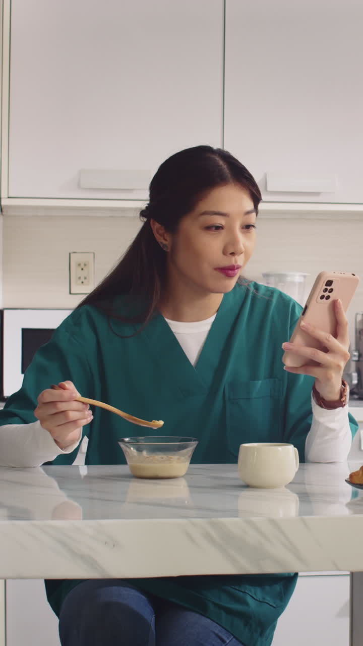 Asian Nurse Eating Yogurt and Using Phone at Kitchen