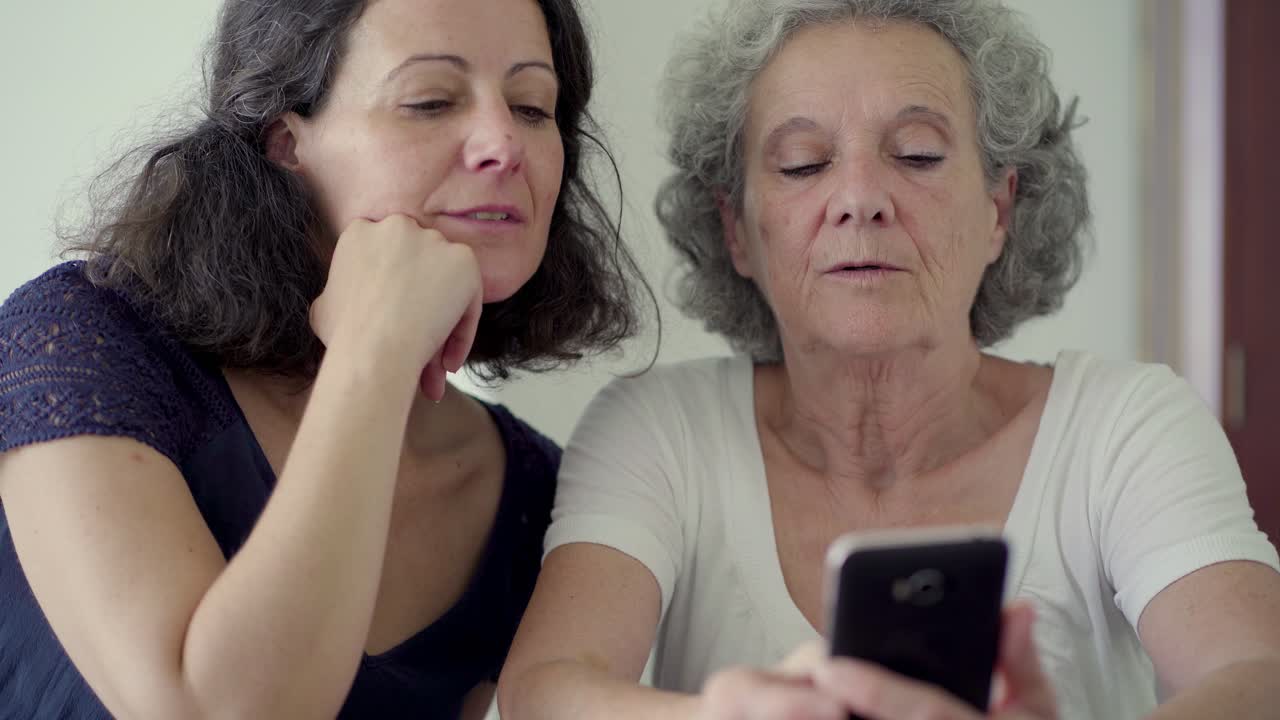 damas maduras sonrientes sentadas a la mesa y usando teléfonos inteligentes.