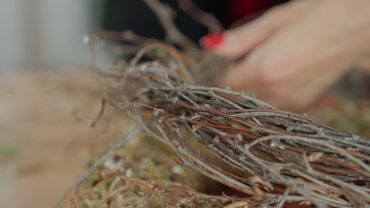 Female florist hands with red nails carefully arranging twigs on straw base to form rustic wreath, highlighting craftsmanship, patience, seasonal creativity, natural decoration process and handmade artistry