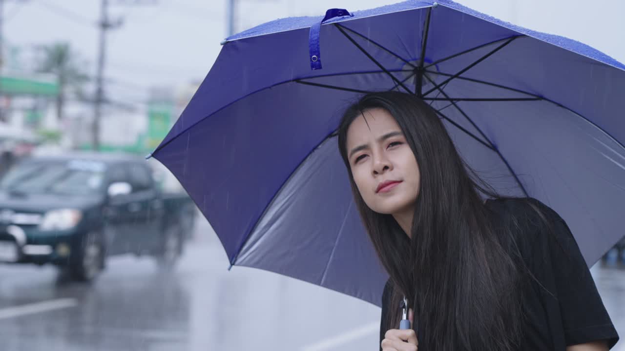 긴 검은 머리카락을 가진 젊은 아시아 여성은 버스 또는 택시를 위해 길을 바라보고, 대중교통 서비스, 비가 오는 계절에 사는 아시아, 열대 기후 변화, 손으로 비방울을 수집합니다.