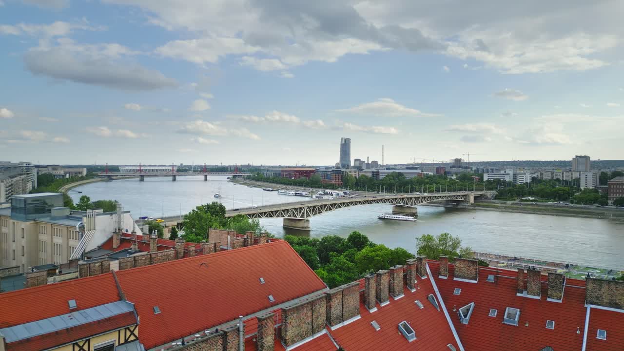 Aerial shot of Budapest, showcasing the Danube River, a busy bridge with moving cars, a boat passing below, terracotta roofs, and lush greenery.