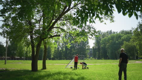 a man ties his shoelaces close to a goalpost with two balls nearby, while his son stands and watches, an elderly man stands in the background