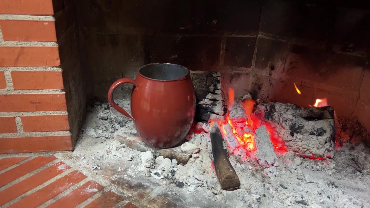 Sebuah tangan meletakkan mug merah di atas kayu bakar yang membara di perapian bata. Pemandangan ini menangkap kehangatan dan pesona pedesaan, cocok untuk proyek bertema musim dingin atau konten rumah yang nyaman.