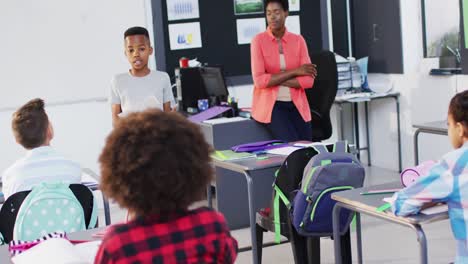 Diverse-female-teacher-and-happy-schoolchildren-at-desks-reciting-in-school-classroom