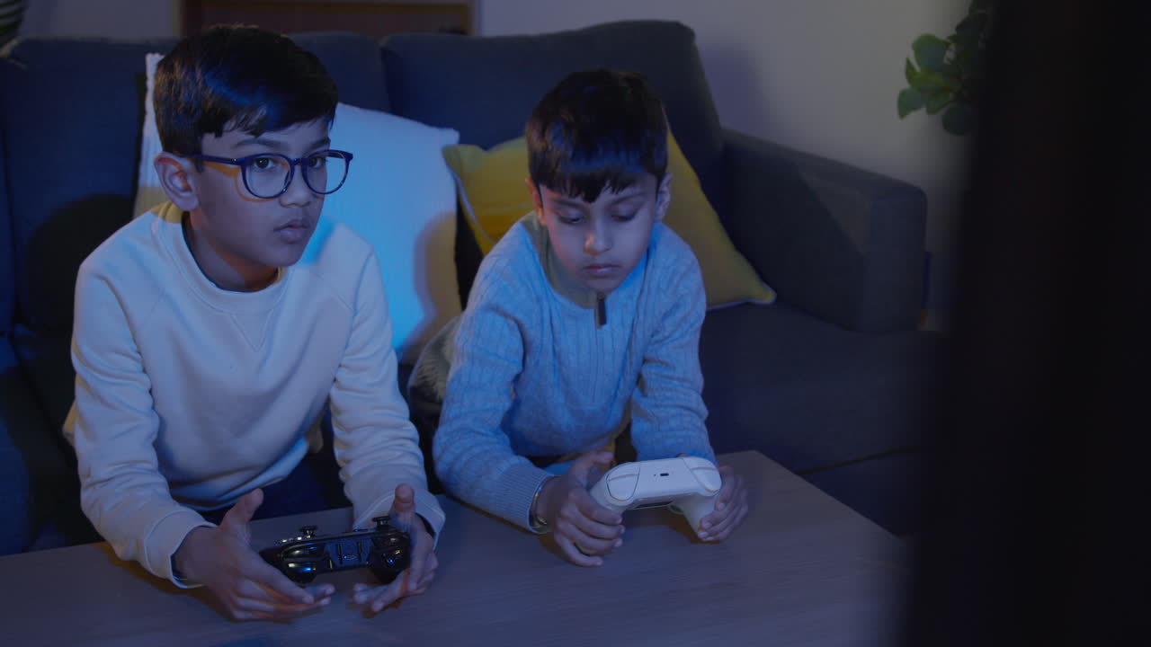 Two Young Boys At Home Playing With Computer Games Console On TV Holding Controllers Late At Night 3