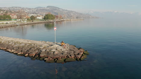 Vista-De-ángulo-Alto-De-La-Mujer-Sentada-Al-Final-Del-Muelle-Con-Vistas-A-Un-Hermoso-Lago-Grande