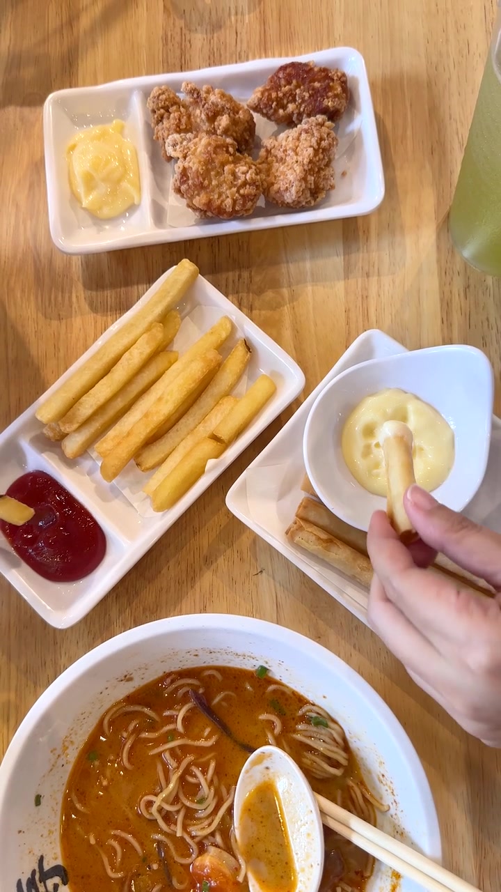 Bàn tay nhúng khoai tây chiên vào nước sốt, xung quanh là mì ramen, gà rán và gia vị, nhìn từ trên cao xuống.