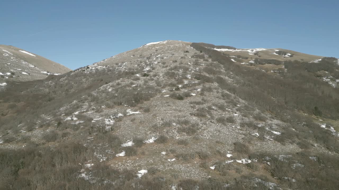 Drone view over Valsorda, Gualdo Tadino, on a sunny winter day. Snow-dusted peaks rise above a vibrant landscape, blending crisp white and rich earthy tones under a clear blue sky