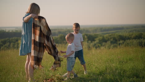 a mother in a blue dress carefully folds a scarf while her two young sons, both wearing white tops, stand close by in a grassy field. the tranquil outdoor setting captures a peaceful family moment