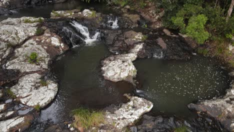 Slow-reveal-of-a-rainforest-creek-flowing-down-towards-a-waterfall-cascading-into-a-deep-tropical-rock-pool-below