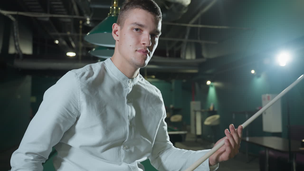 색 셔츠와 회색 바지를 입은 남자가 희미하게 밝은 빌리어드 홀에서 녹색 술집 테이블에 앉아 있으며, 진지한 표정과 집중된 시선을 가진 큐 스틱을 들고 있습니다. 상공 조명은 극적인 그림자를 던집니다.