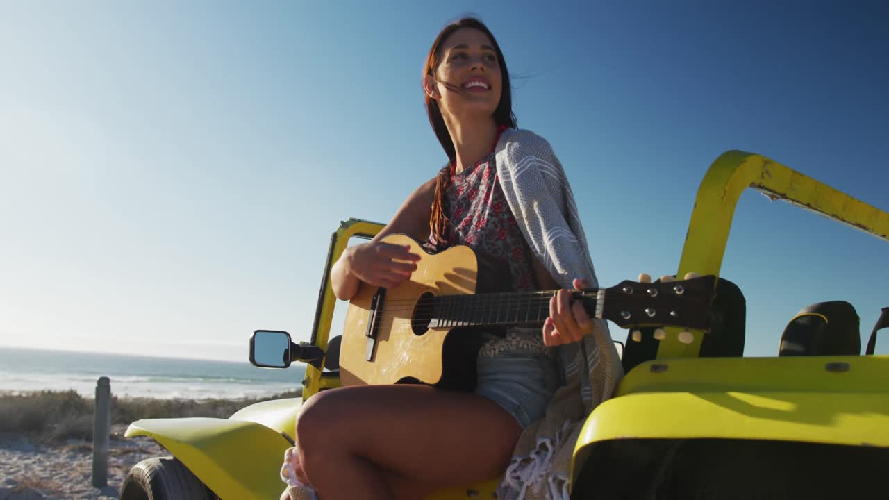 feliz mujer caucásica sentada en un buggy de playa junto al mar tocando la guitarra
