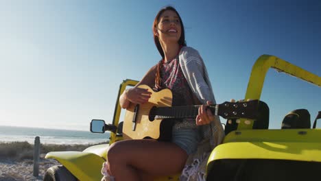 happy caucasian woman sitting in beach buggy by the sea playing guitar