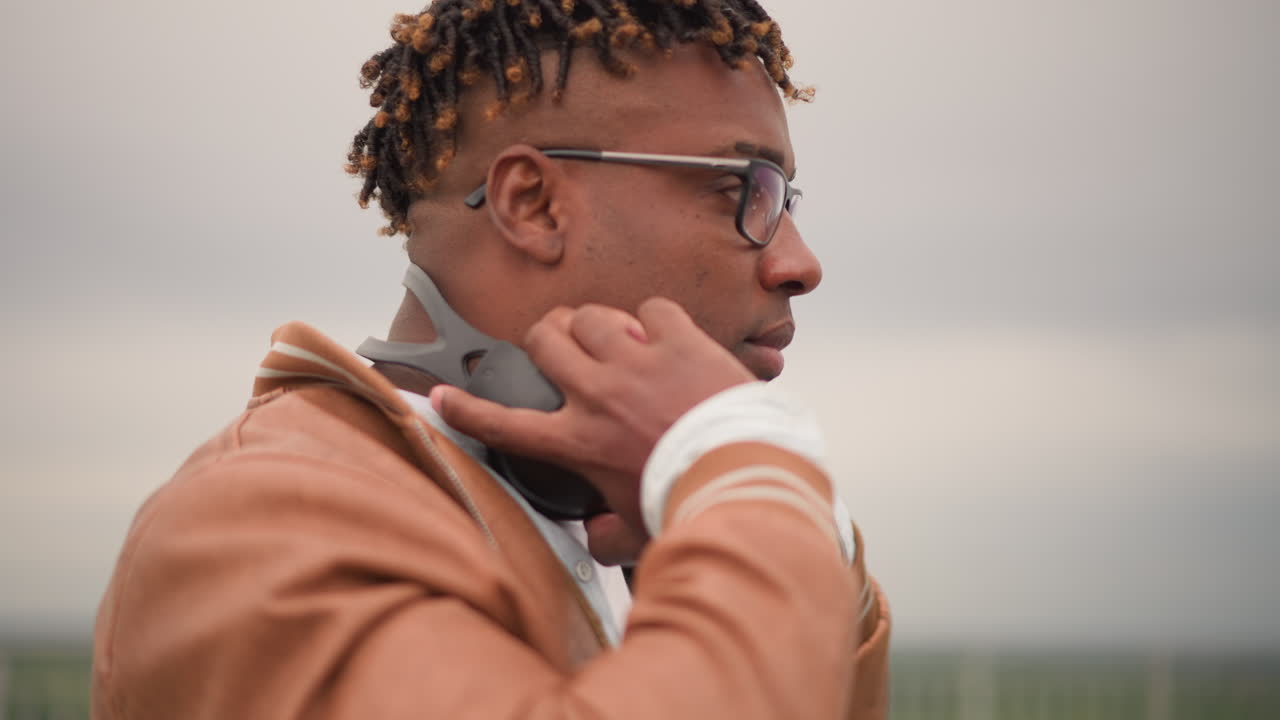 Rooftop DJ wearing headphones adjusting headset, scanning city skyline, testing beats and ambient levels, calm contemplative expression, leather jacket and glasses, cable draped, dusk mood, preparing