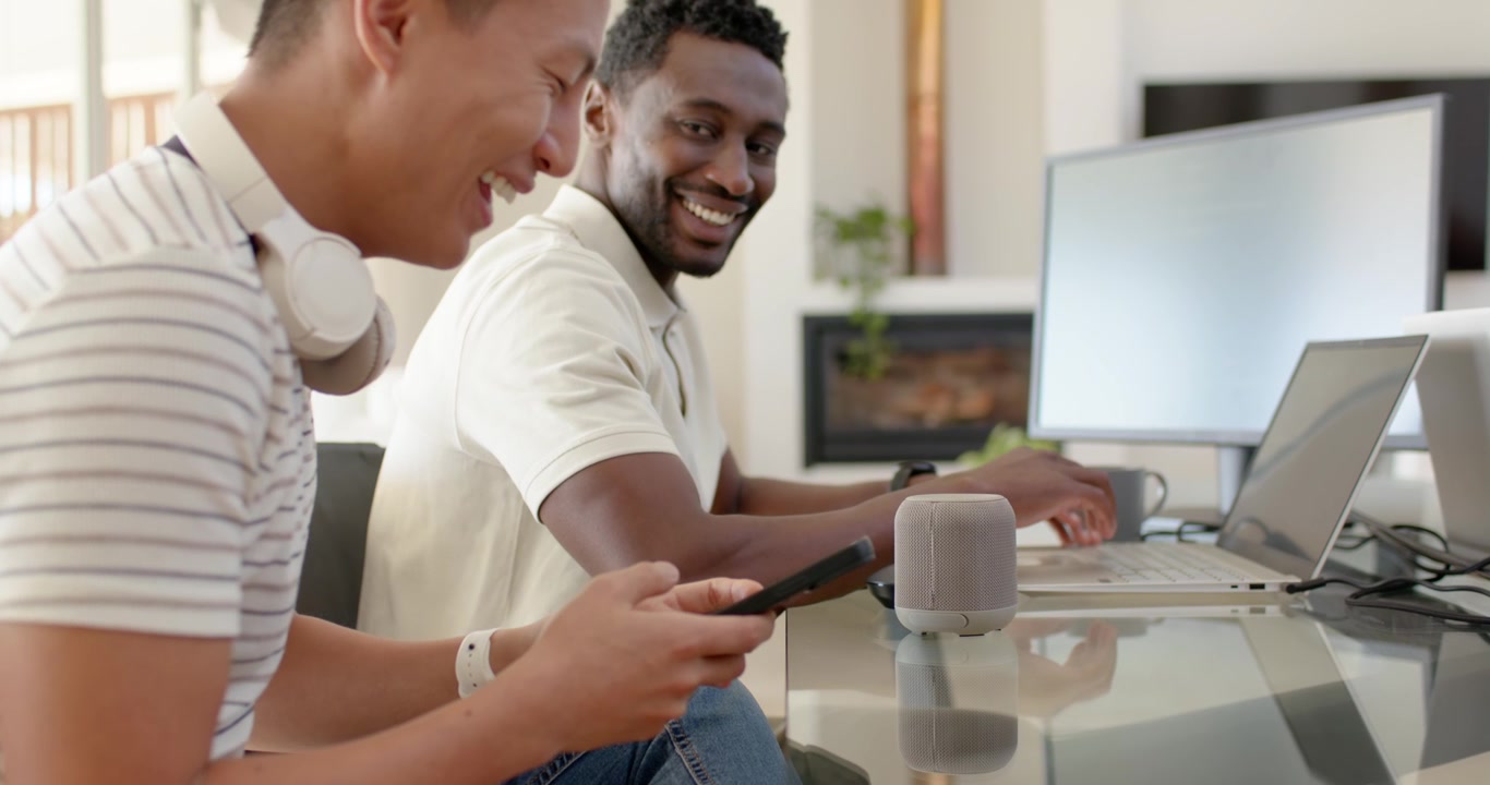 Diverse men smiling as left tapping phone right using speaker typing laptop at home office working