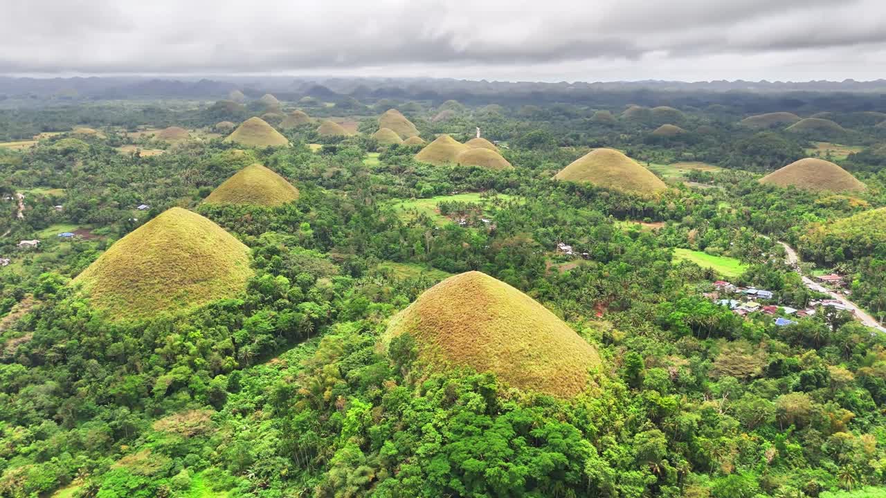 Aerial view of the Chocolate Hills in Bohol, Philippines, featuring conical grass-covered mounds rising from dense green forest, with hundreds of hills stretching across the horizon under cloudy skies