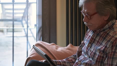 Old-Man-Wih-Gray-Hair-Watching-On-A-Tablet-While-Sitting-On-The-Couch