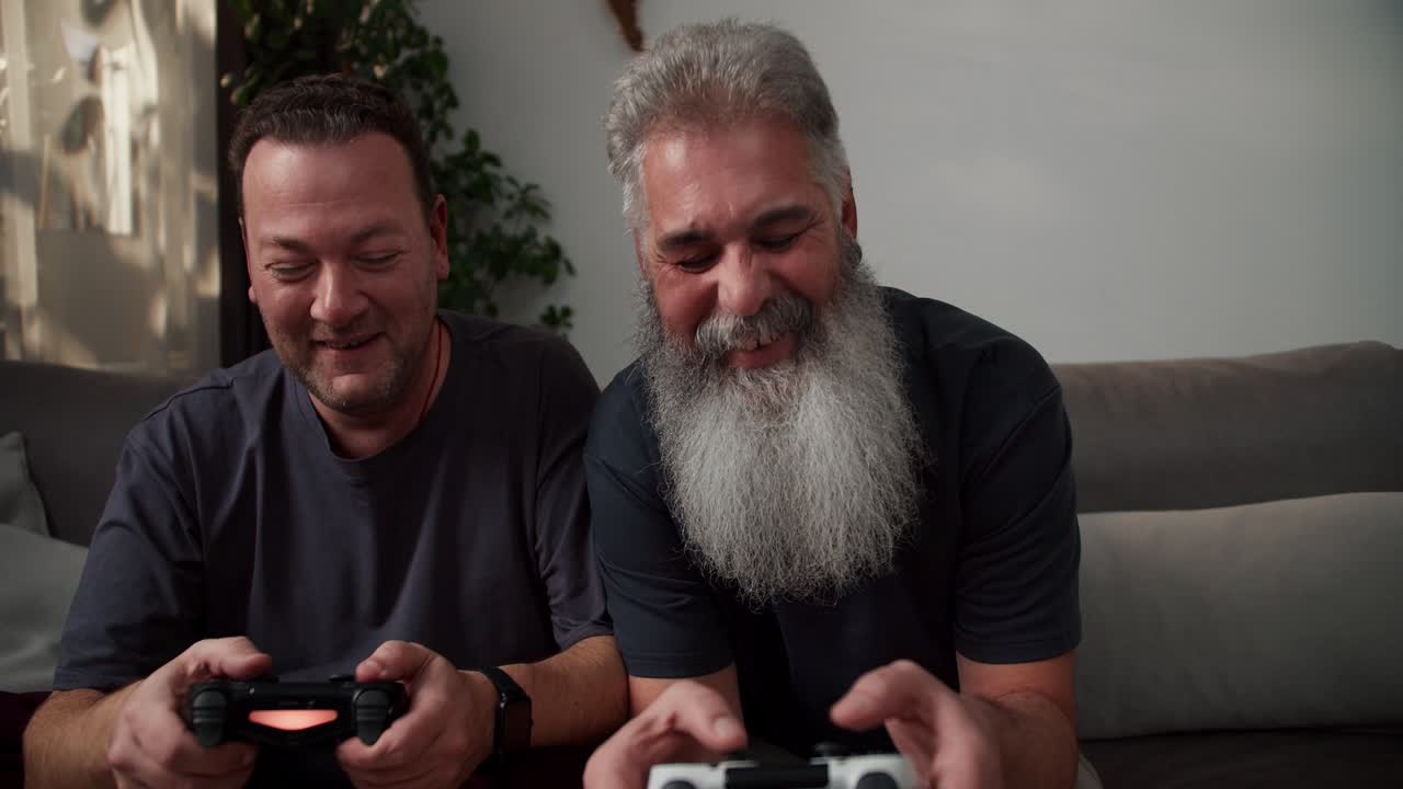 ritratto di un uomo felice con i capelli grigi e una barba grigia lussureggiante che gioca a videogiochi usando un joystick insieme al suo fidanzato brunetto con la paglia in una maglietta grigia seduto sul divano in un appartamento moderno la sera