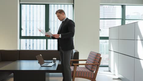 businessman in a dark suit stands reviewing documents in a modern office space featuring floor-to-ceiling windows, a laptop on a dark desk, a patterned accent chair, and minimalist white storage units