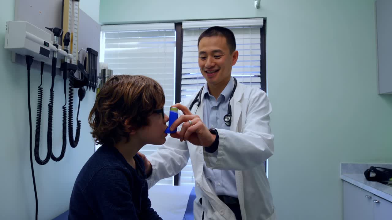 Young asian male doctor helping a caucasian boy patient in using asthma inhaler in clinic 4k